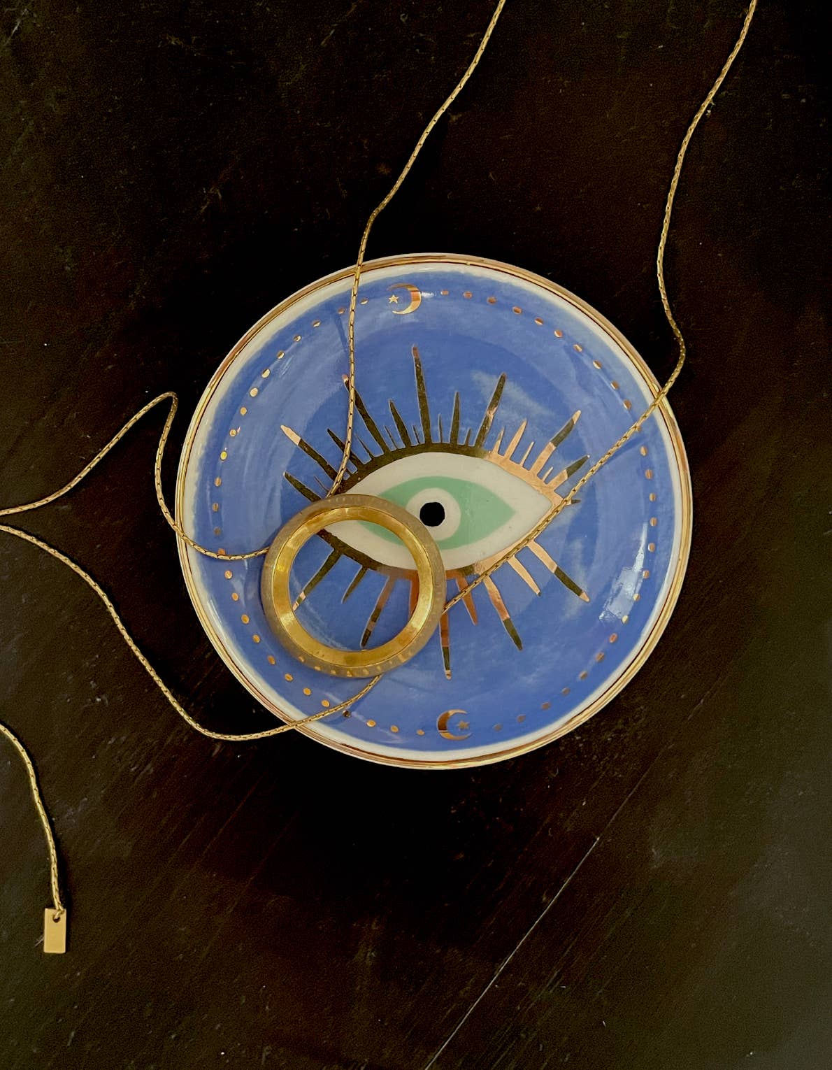 An Idlewild Co. Porcelain Trinket Dish - Evil Eye featuring a round, gold-edged ceramic design with gold rays, holding a gold ring and a thin gold chain.