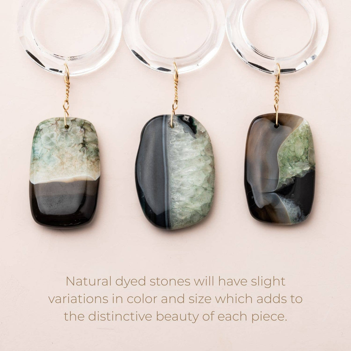 Three rectangular, multicolored “Stone Suncatcher - Sun Rays/Black/Blue Agate” keychains by Scout Curated Wears, each adorned with a gold ring, are displayed in a row above a beige background featuring text about natural dyed stone variations—reminiscent of elegant stone suncatchers.