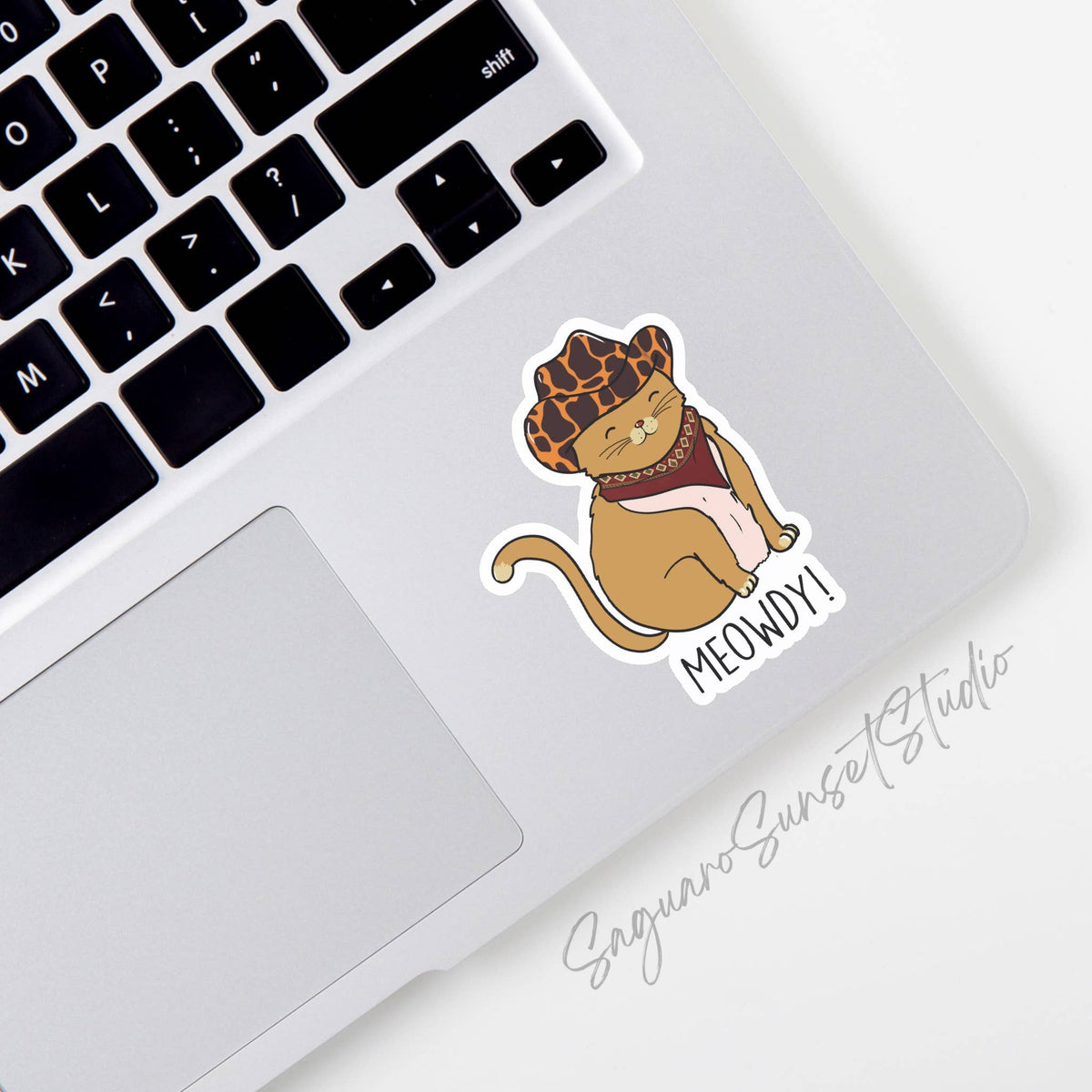 Add a touch of fun to your laptop with the Meowdy Cowboy Cat Sticker by Saguaro Sunset Studio—a vinyl sticker featuring a smiling cartoon cat in a cowboy hat and bandana, next to the word “MEOWDY!” in the bottom right corner of your keyboard.