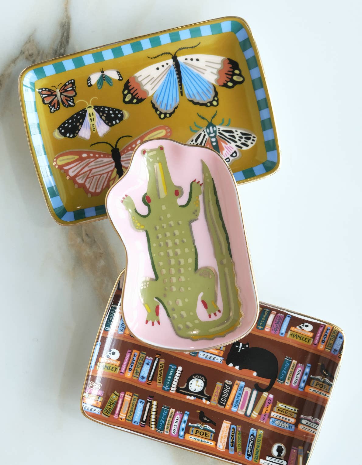 Three stacked hand-painted ceramic dishes: a rectangular tray with colorful moths on mustard, a narrow tray with a green crocodile on pink, and a Porcelain Trinket Tray - Bookshelves Library.