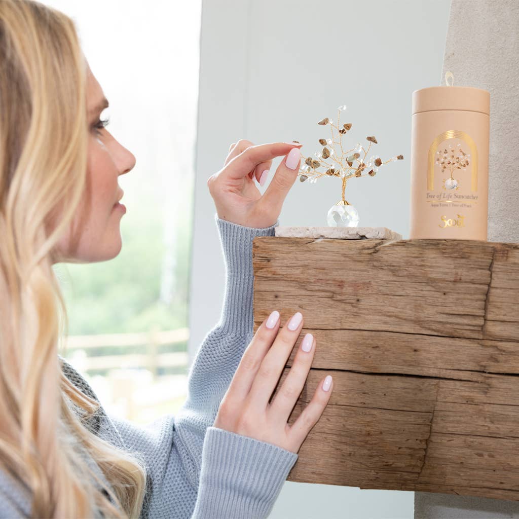 A woman in a blue sweater places a small Suncatcher Tree of Life - Turquoise/Tranquility by Scout Curated Wears with gemstone leaves on a rustic wooden shelf.