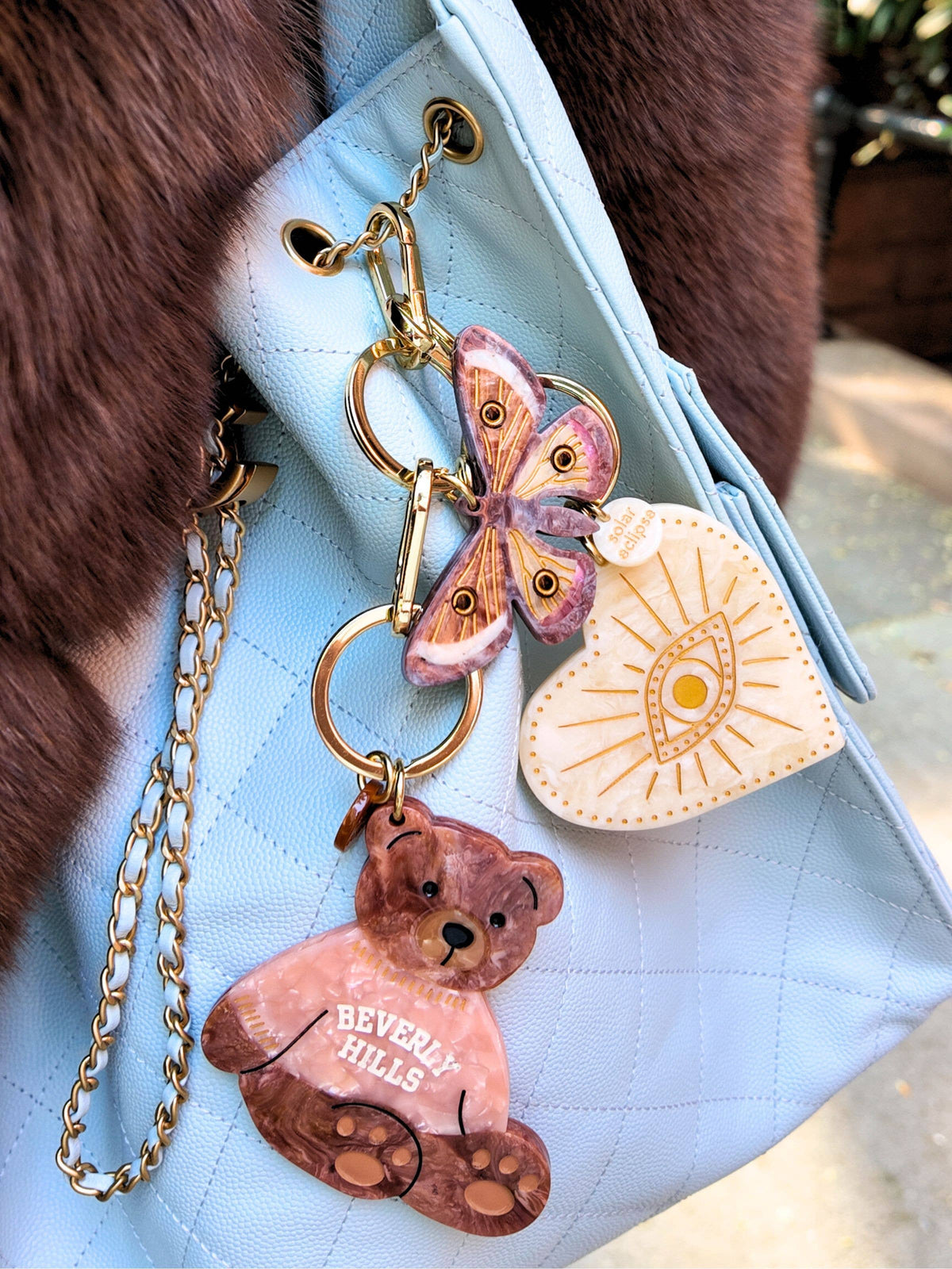 A light blue quilted bag adorned with the Solar Eclipse Celestial Heart Bag Charm + Keychain, featuring three keychains: a brown teddy bear labeled Beverly Hills, a pink and gold butterfly, and a white heart with a gold evil eye design.