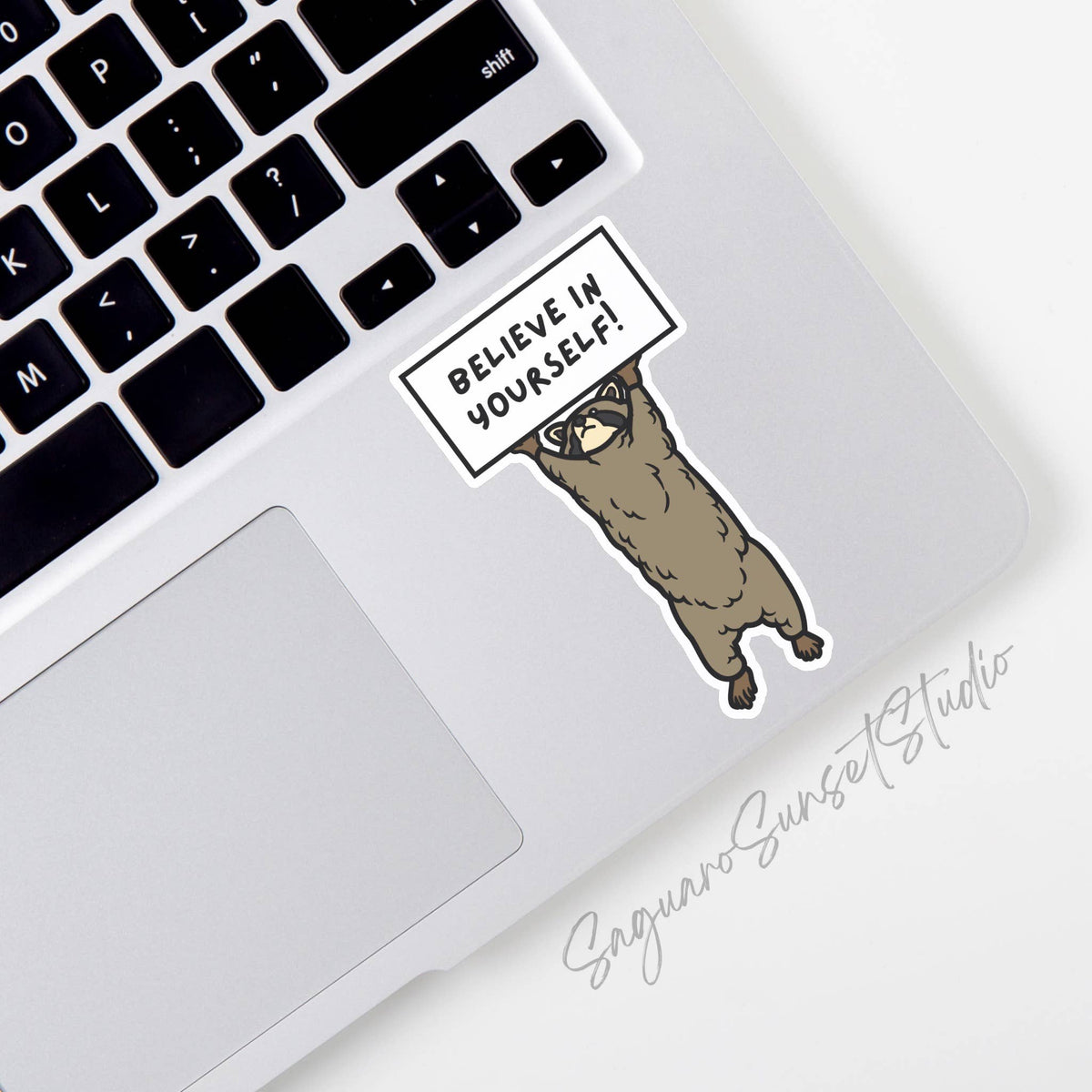 A waterproof Believe In Yourself! Raccoon Sticker by Saguaro Sunset Studio is placed near the keyboard on a silver laptop with Saguaro Sunset Studio in script on the laptop’s surface.