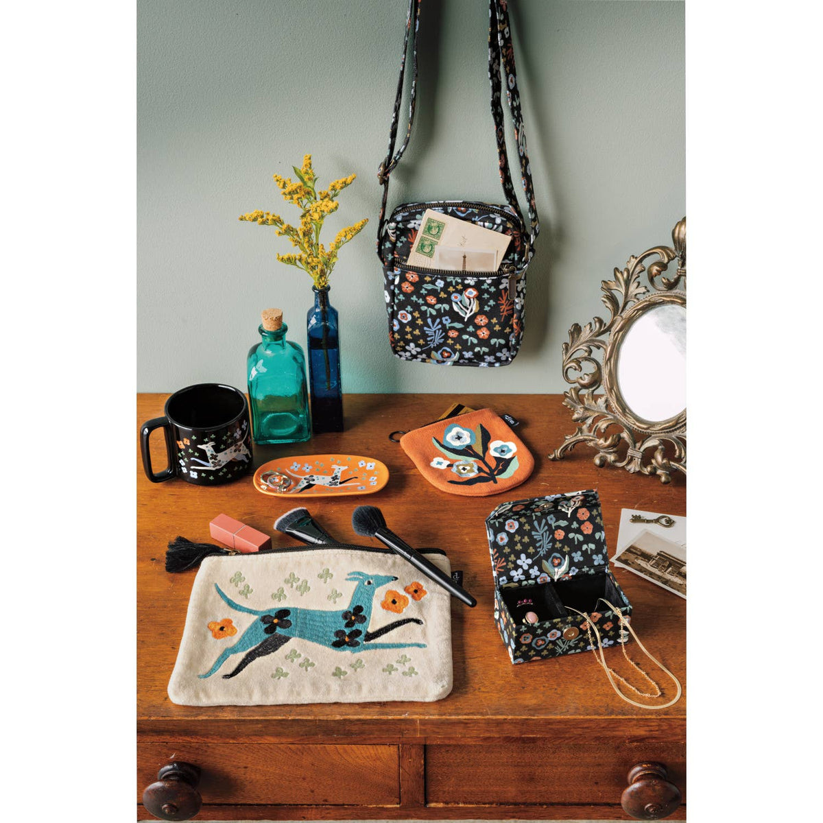 A Dogwood Cotton Crossbody Bag - Multi Floral/Black with an adjustable strap hanging above a wooden desk scattered with matching accessories, including a zippered pouch, a small jewelry box, and a ceramic tray.