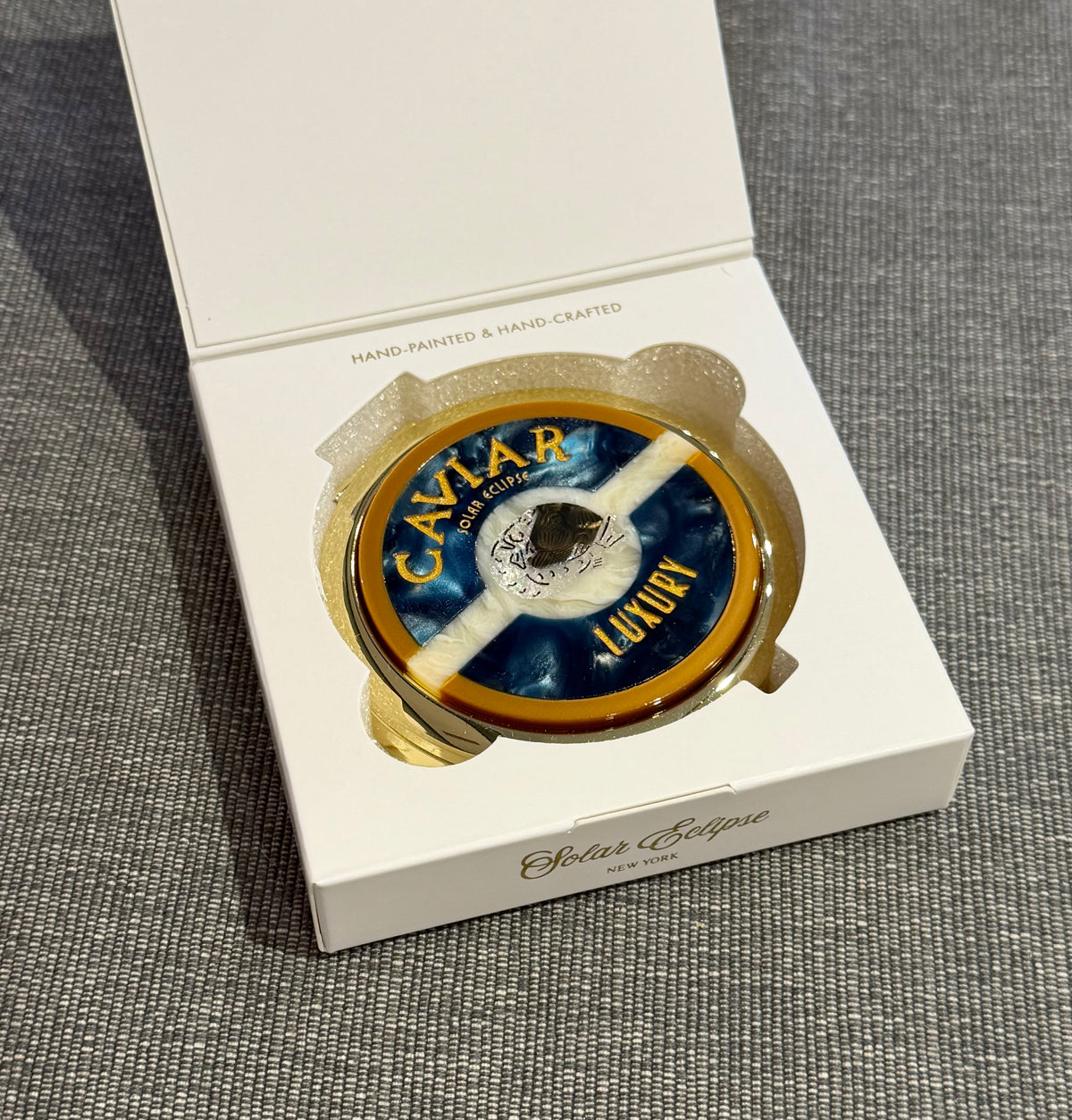Solar Eclipse Caviar Handbag Hero Pill Compact - Hand-Painted, a round marble-patterned case with a blue and white design and gold Caviar Luxury lettering, nestled inside an open white gift box.