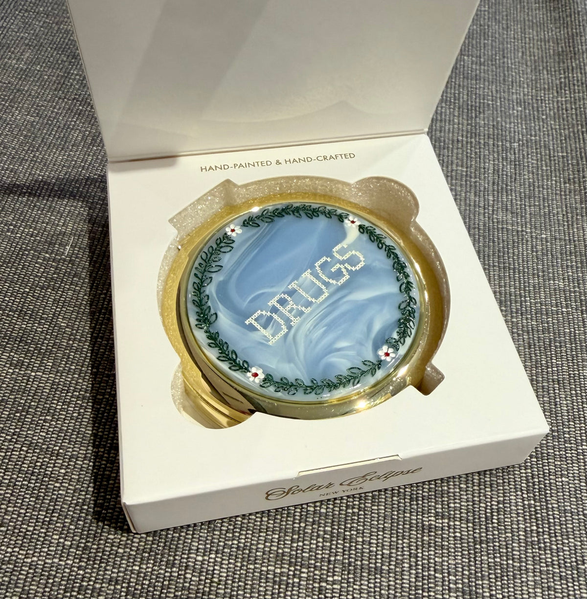 A round gold Solar Eclipse Cross-Stitch Drugs Handbag Hero Pill Compact - Hand - Painted with a blue marbled lid featuring the word DRUGS in white stitched letters, framed by a floral wreath, inside an open white gift box.