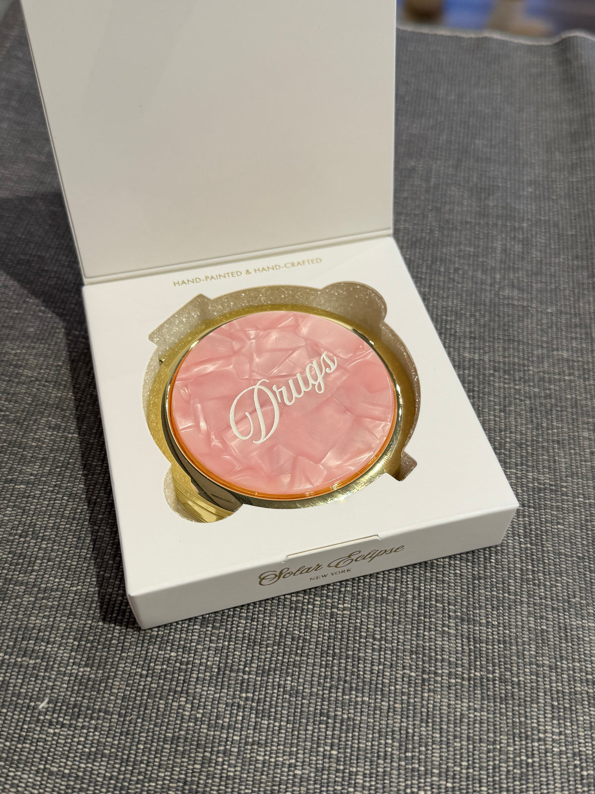 A round, pink, pill-shaped Solar Eclipse Pink Drugs Handbag Hero Pill Compact - Hand - Painted, featuring the word Drugs in script, displayed in its open white product box as a portable handbag pill organizer.
