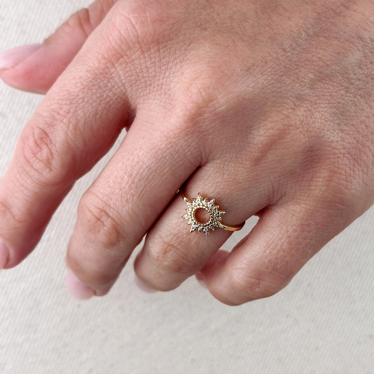 A hand adorned with the GoldFi Gold Filled CZ Open Starburst Ring—an open starburst design embellished with sparkling clear stones—graces the ring finger against a softly textured, light backdrop.
