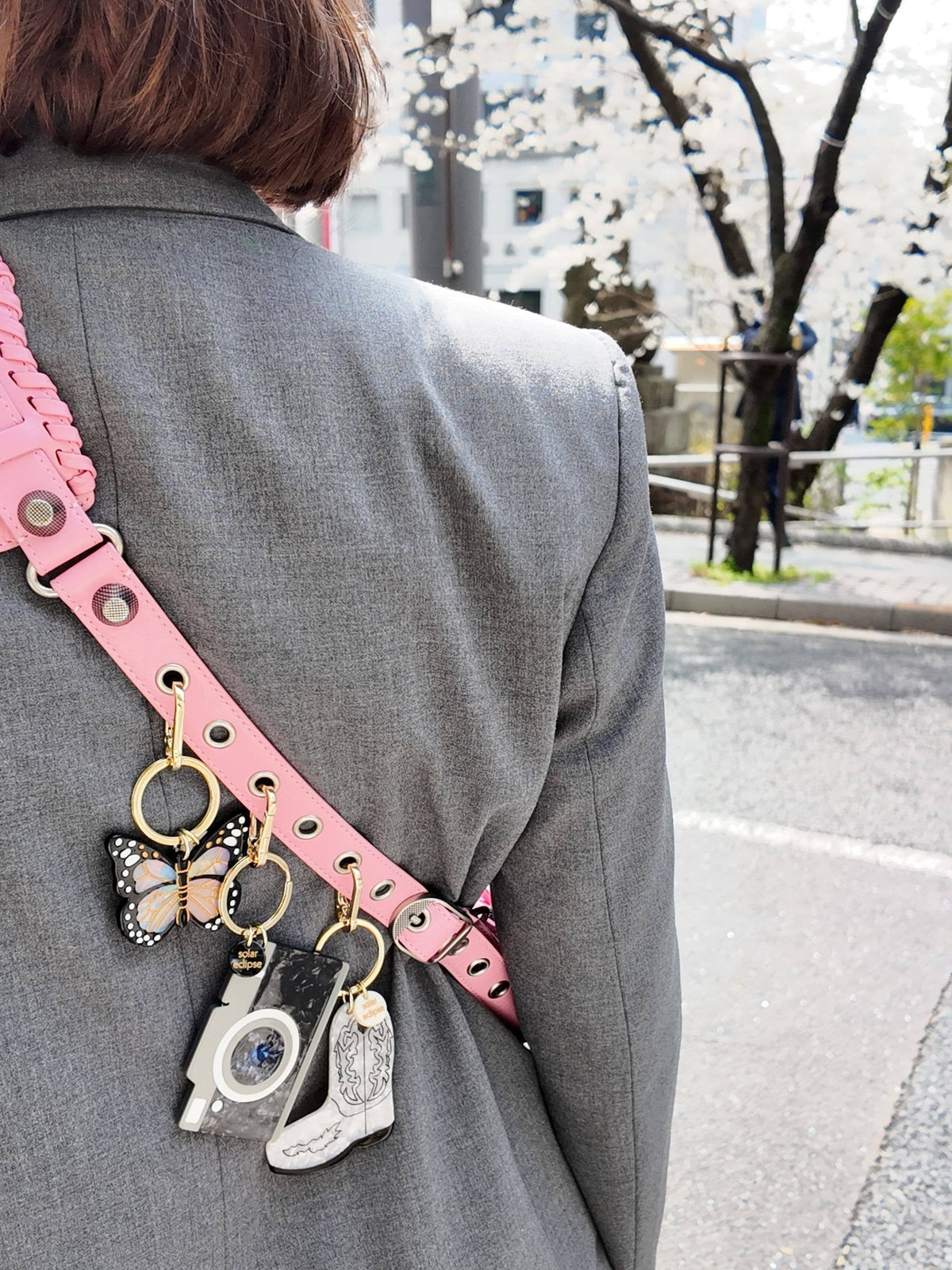 A person in a gray blazer with a pink strap over their shoulder, displaying hand-painted keyrings—including a butterfly, a white camera, and a small silver object—stands outdoors near blossoming trees; among the accessories is the Solar Eclipse Monarch Butterfly Bag Charm + Keychain, crafted from biodegradable acetate.