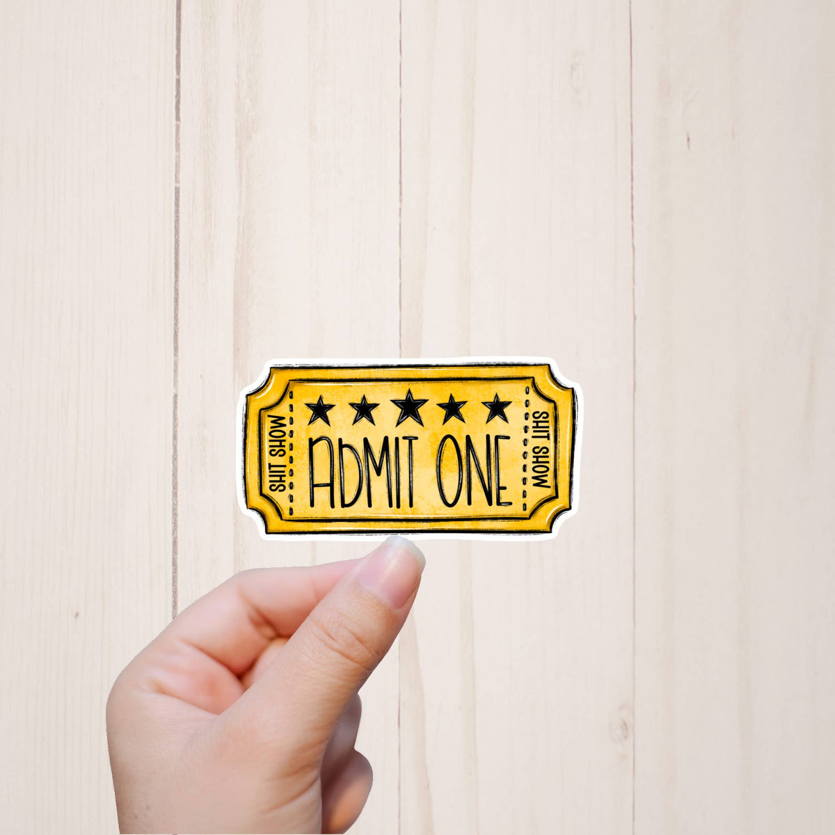 A hand holds a Shit Show Sticker by Sorry Not Sorry Co, styled like a yellow Admit One ticket (3″ x 1.4″) with black stars, against a light wooden background. Durable vinyl sticker.
