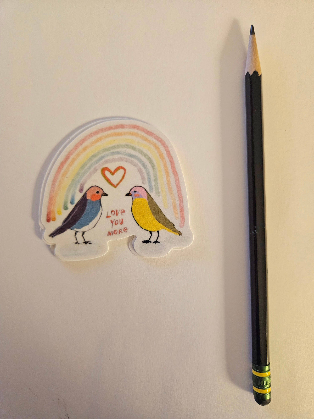 The Birds Rainbow Love You More Sticker by Blueberry Road Cards shows two colorful birds under a rainbow and red heart, with “LOVE YOU MORE” between them, next to a black pencil with green eraser on a white background.