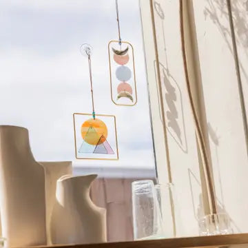 Two Moon Phase Stained Glass Light Catchers by Scout Curated Wears hang in a sunlit window next to minimalist ceramic vases and glassware, casting colorful reflections and shadows on the wall behind them.