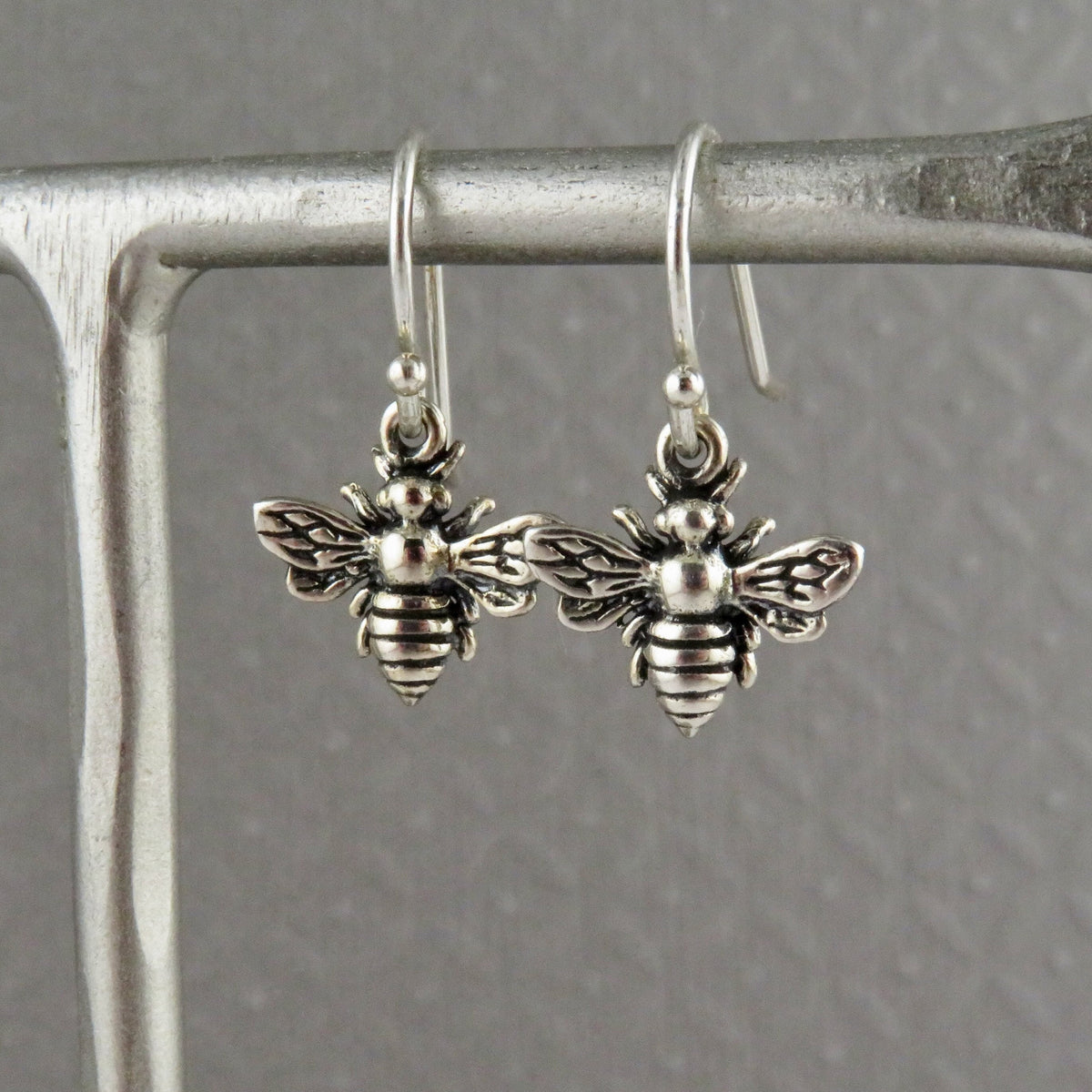 A pair of silver bee-shaped dangle earrings hang gracefully from a metal stand, complemented by a textured gray background—an match for the Nina Designs Small Bee Necklace in sterling silver.