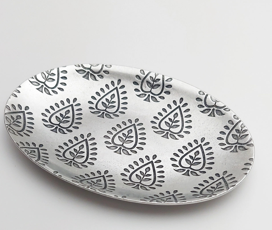 An oval silver dish with a repeated black leaf pattern etched into the surface, set against a plain white background.