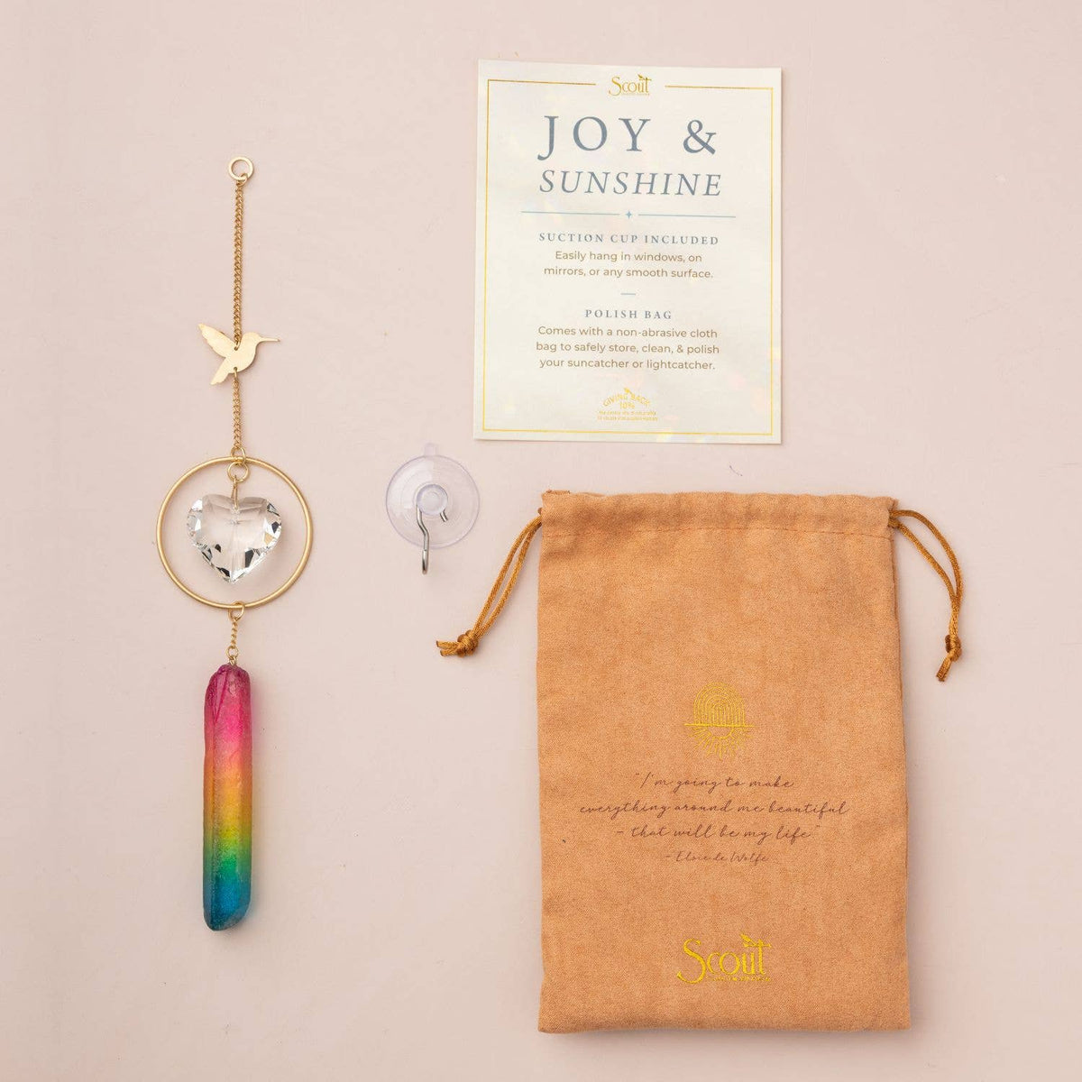 The Scout Curated Wears Stone Suncatcher - Hummingbird/Rainbow Stone, featuring a rainbow crystal, delicate natural stone accents, a golden bird charm, a suction cup, an instruction card, and a tan drawstring pouch with gold text, is neatly arranged on a light peach background.