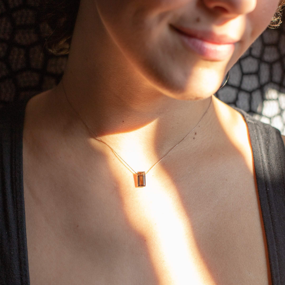 A person smiles softly, sunlight and shadows playing across their neck, as they wear the Smoky Quartz Refined Necklace for Release by SoulKu with its slender rectangular pendant.
