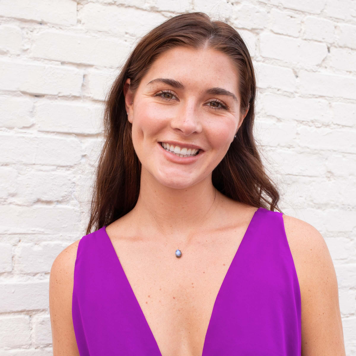 A woman with long brown hair smiles at the camera, wearing a sleeveless purple top and the SoulKu Labradorite Luxe Necklace for Transformation, standing in front of a white brick wall.