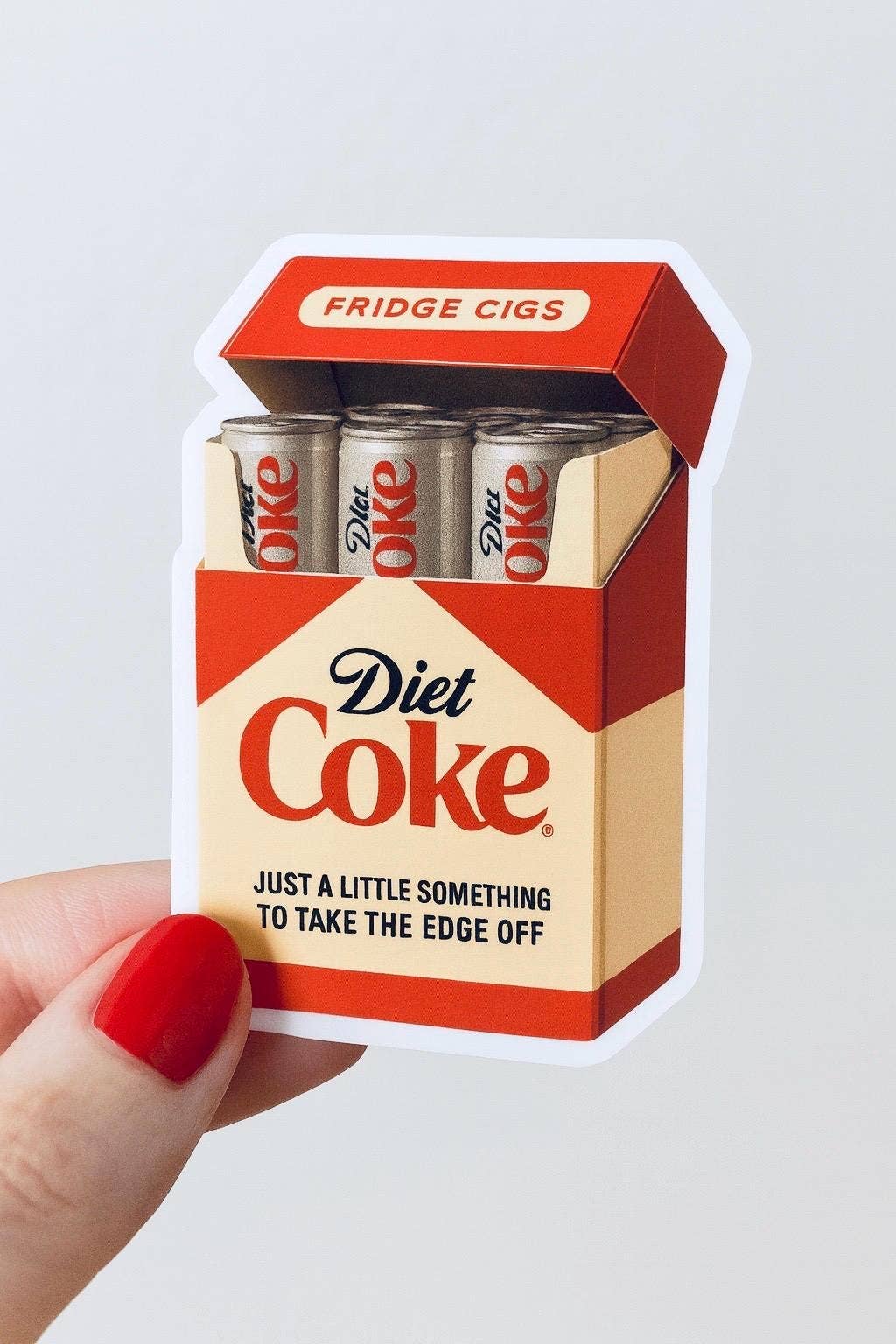 A hand with red nail polish holds the Diet Coke Fridge Cigs Sticker by Hello Brand | Sticker My Art, featuring a weather-resistant vinyl design of a cigarette pack filled with Diet Coke cans and cheeky text.