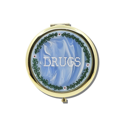 Round gold Cross-Stitch Drugs Handbag Hero Pill Compact - Hand - Painted featuring a blue marbled lid with the word DRUGS in white cross-stitch lettering, surrounded by a painted green vine and flower border.