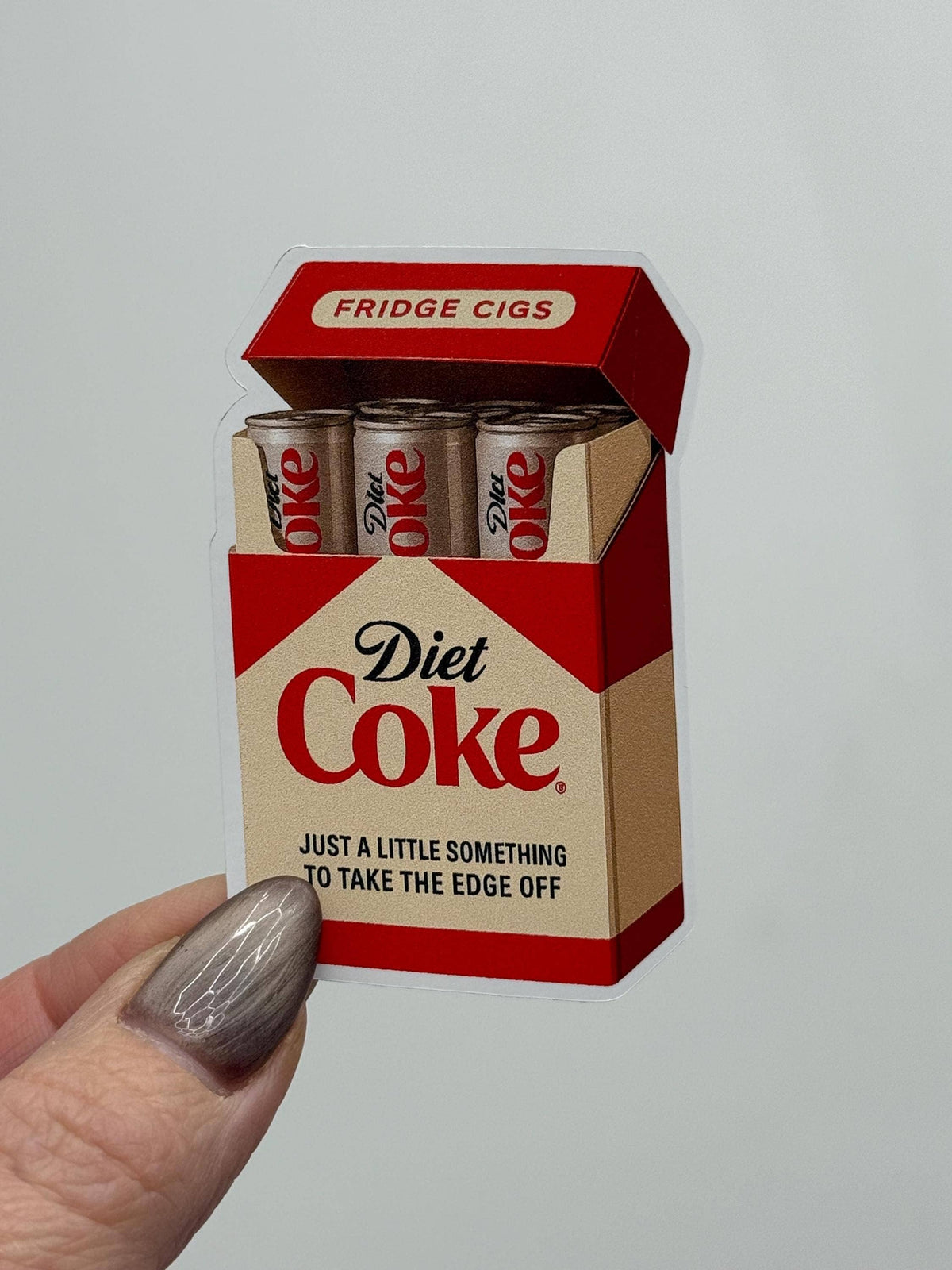 A hand holds a weather-resistant Diet Coke Fridge Cigs Sticker by Hello Brand | Sticker My Art, designed like a cigarette pack with cans and the phrases “Diet Coke,” “Just a little something to take the edge off,” and “Fridge Cigs.”.