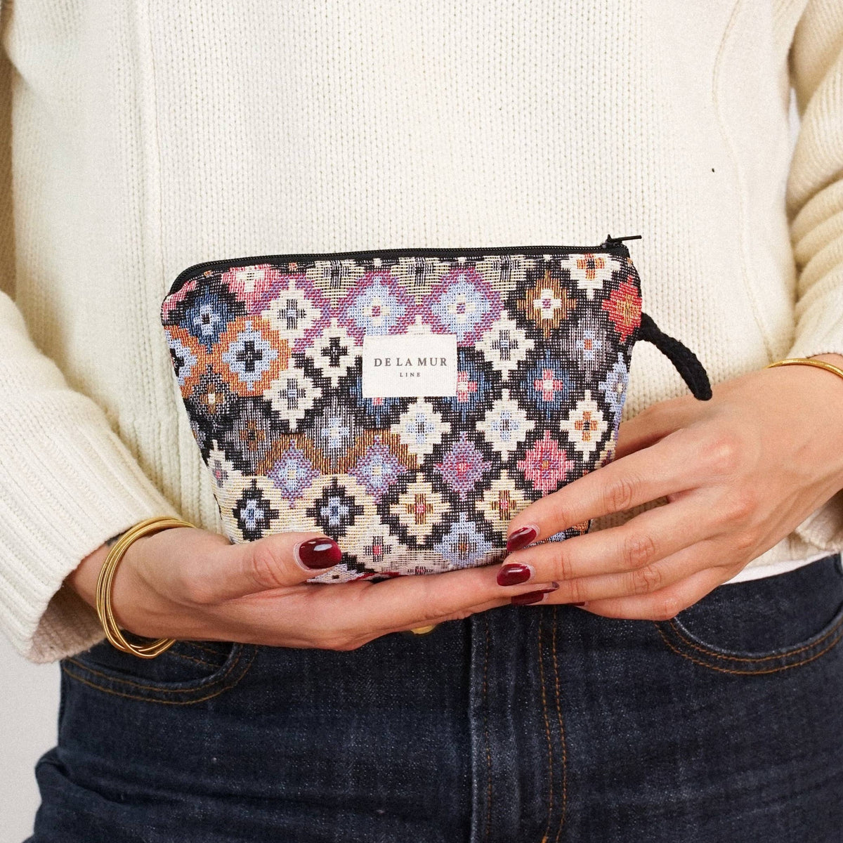 A person in a cream sweater and dark jeans holds the DE LA MUR LINE US Inc Geometric Jacquard Small Makeup Bag – Multicolor (“Ore”) in both hands at waist level. Only their torso and hands are visible.