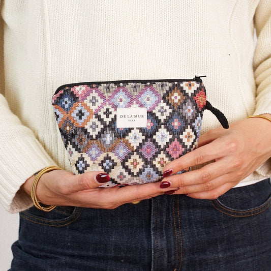 A person in a cream sweater and dark jeans holds the DE LA MUR LINE US Inc Geometric Jacquard Small Makeup Bag – Multicolor (“Ore”) in both hands at waist level. Only their torso and hands are visible.
