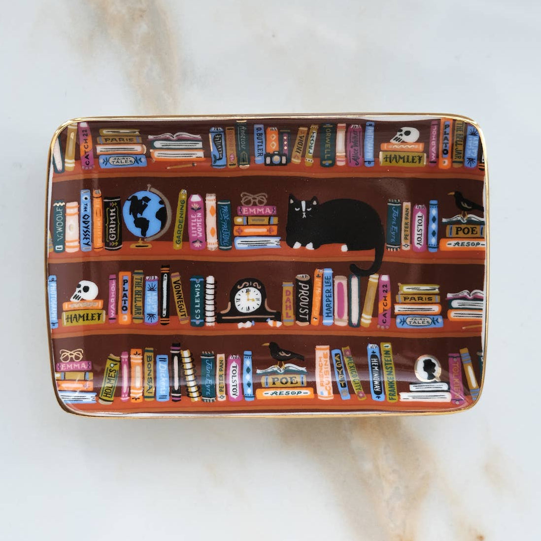 Idlewild Co. Porcelain Trinket Tray - Bookshelves Library, a rectangular tray with a gold rim featuring a hand-painted illustration of bookshelves, a black cat, a globe, a clock, and a bird.