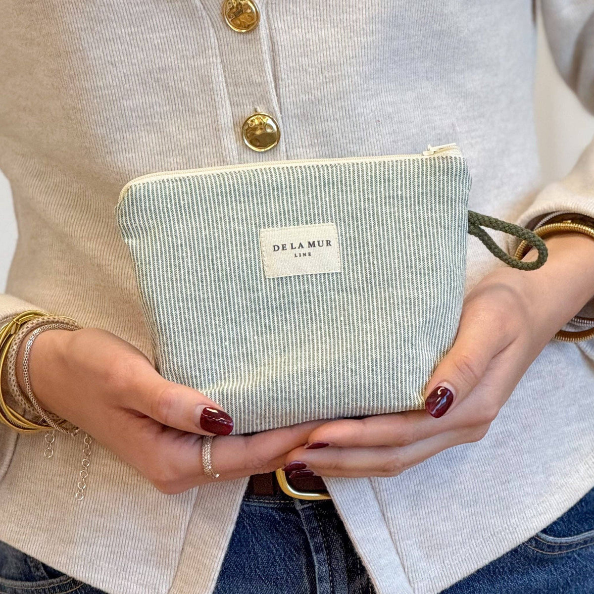 A person wearing a beige cardigan and jeans holds the Striped Small Makeup Bag – Light Green & Green (Teo) by DE LA MUR LINE US Inc, featuring a DE LA MUR label and crafted from elegant jacquard fabric. With bracelets adorning both wrists and neatly manicured nails, they showcase this stylish makeup bag, which is proudly made in Barcelona.