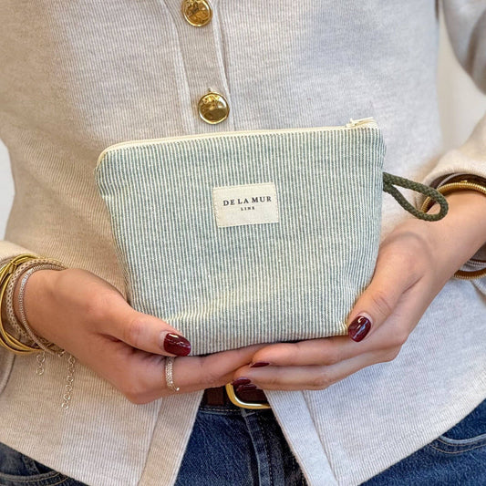 A person wearing a beige cardigan and jeans holds the Striped Small Makeup Bag – Light Green & Green (Teo) by DE LA MUR LINE US Inc, featuring a DE LA MUR label and crafted from elegant jacquard fabric. With bracelets adorning both wrists and neatly manicured nails, they showcase this stylish makeup bag, which is proudly made in Barcelona.