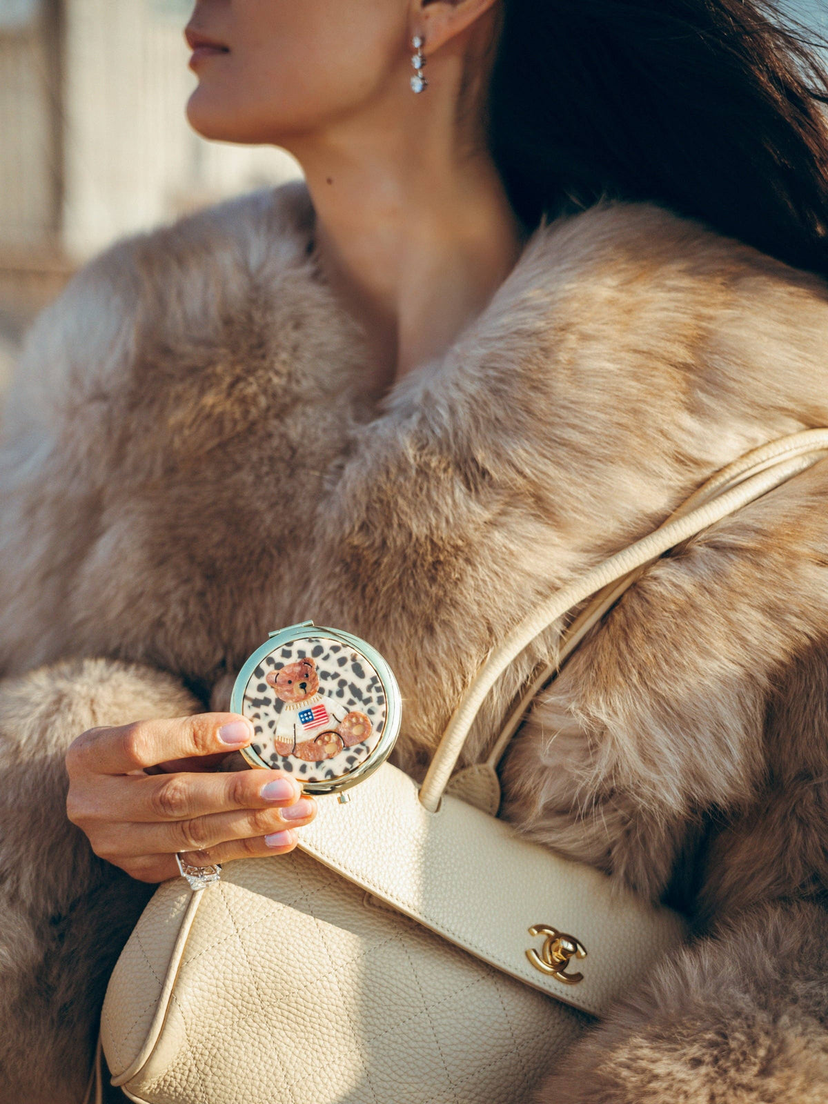 A person in a fur coat holds a small, round Teddy Bear Americana Handbag Hero Pill Compact - Hand-Painted featuring a hand-painted teddy bear wearing a sweater with an American flag design.