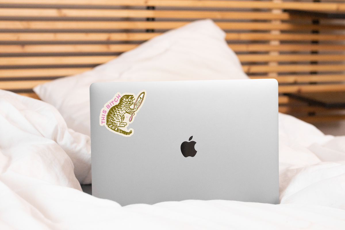 A silver MacBook with a Yarnival This Bitch Cat Sticker, featuring a cat holding a knife and the words THIS BITCH, rests on a white bed with wooden slats in the background.
