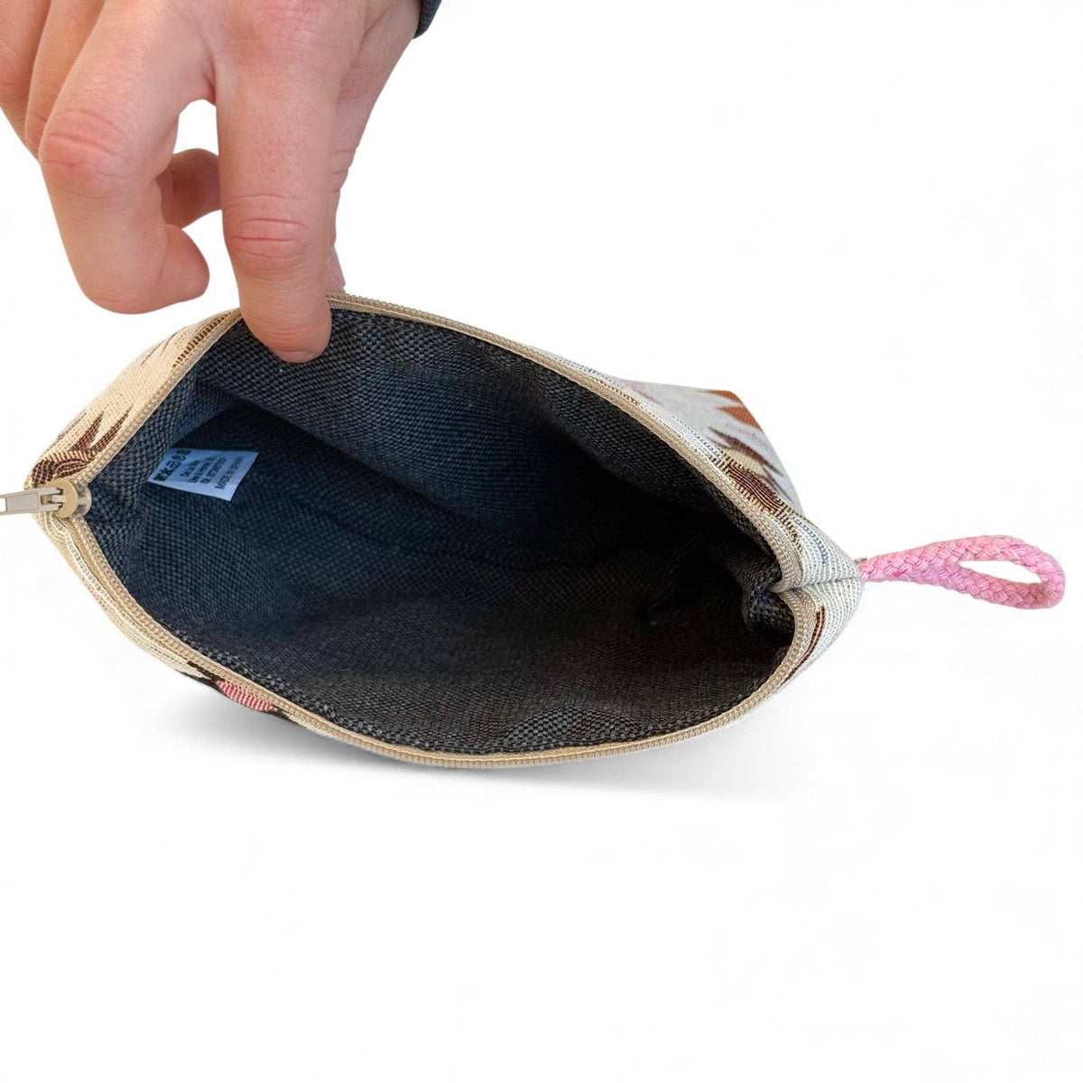 A hand holds open the Geometric Jacquard Small Makeup Bag – Pink & Orange (Royal) by DE LA MUR LINE US Inc, revealing its dark fabric interior and pink woven loop on the side; the pouch features an Aztec-inspired geometric pattern on the exterior and includes a fabric brand tag inside.