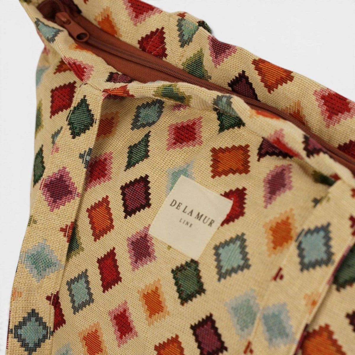 A close-up of the Geometric Jacquard Large Zippered Tote Bag – Multicolor (Zac) from DE LA MUR LINE US Inc shows its vibrant diamond-patterned jacquard fabric and a white label with the brand name, DE LA MUR LINE, sewn at the center. The partially open top zipper reveals the bag’s rich, textured material.