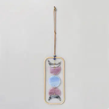 Introducing the Stained Glass Light Catcher - Moon Phase by Scout Curated Wears: This hanging wall décor features a gold oval frame adorned with three gray half-moon shapes and two full circles in pink and blue, arranged vertically to represent the phases of the moon. Finished with a brown string for easy hanging, it beautifully stands out against a light gray background.