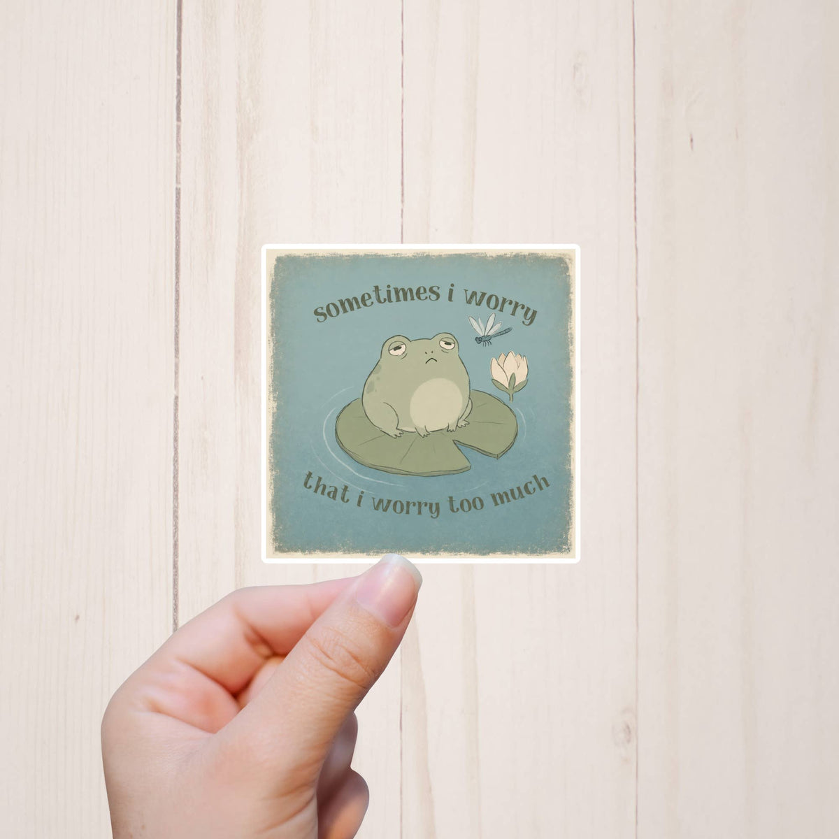 A hand holds the Sometimes I Worry, Funny Frog Sticker by Sorry Not Sorry Co, featuring a sad frog on a lily pad with the text “sometimes I worry that I worry too much.”.