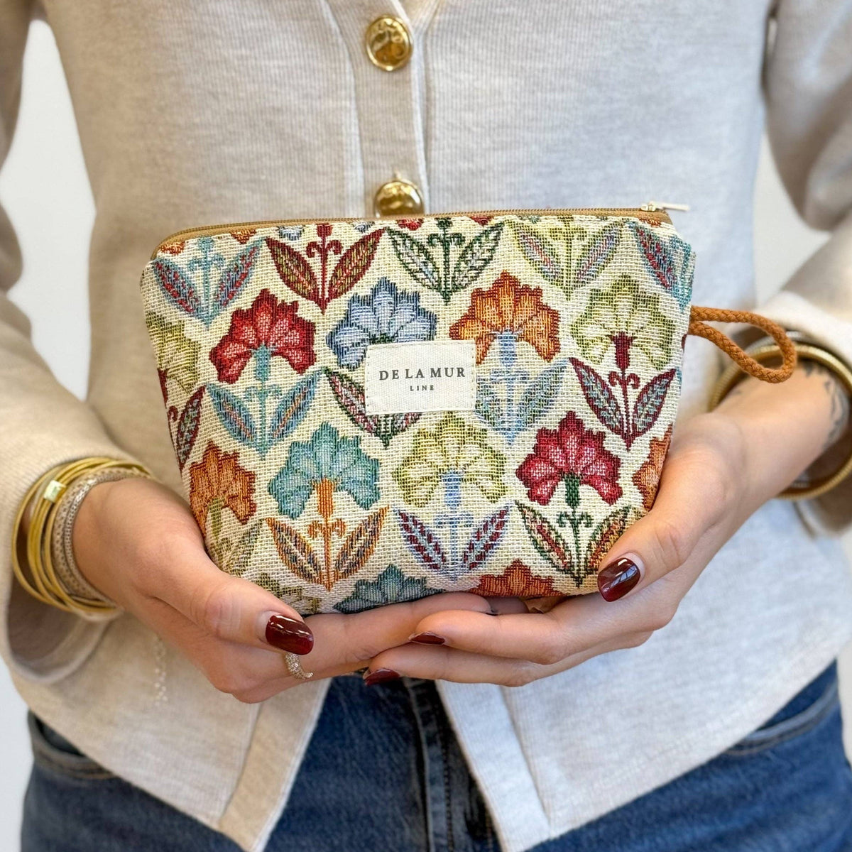 A person in a light cardigan and jeans holds the Floral Jacquard Small Makeup Bag – Multicolor (Flora) by DE LA MUR LINE US Inc. The compact pouch showcases beautifully woven jacquard fabric, while the individual’s painted nails and stacked bracelets add a personal, stylish touch.