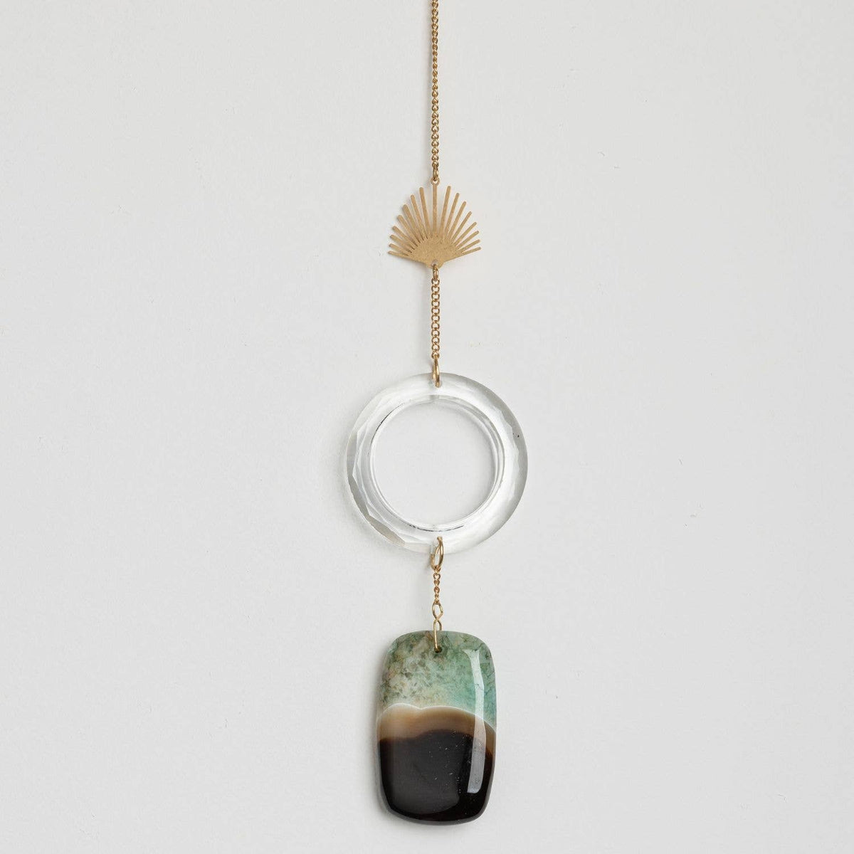 Scout Curated Wears’ “Stone Suncatcher - Sun Rays/Black/Blue Agate” features a gold chain adorned with a fan-shaped sun ray charm, a clear circular ring evoking the look of a sun catcher, and a rectangular pendant striped in green, white, and black.