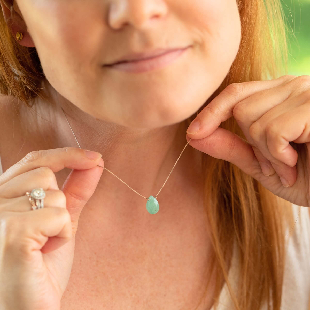 A woman with light skin and auburn hair holds the SoulKu Green Aventurine Gemstone Necklace - Healing, smiling gently, with gemstone jewelry on her left hand against a blurred green background.