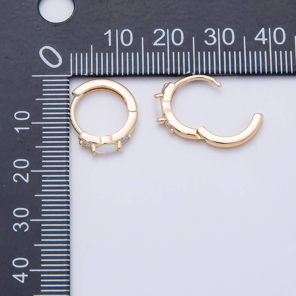 Aim Eternal Gold Filled 13mm White Opal/CZ Huggie Hoops, one featuring a central white opal and clear cubic zirconia and the other an open design, resting beside a black ruler.