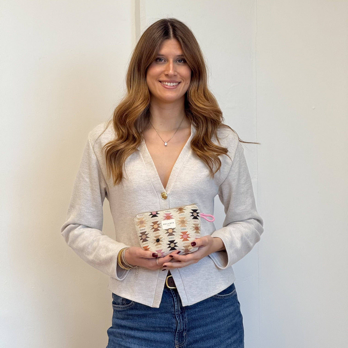 A woman with long wavy hair, dressed in a light-colored cardigan and blue jeans, stands smiling against a plain wall while holding the DE LA MUR LINE US Inc Geometric Jacquard Small Makeup Bag – Pink & Orange (Royal) in both hands in front of her.