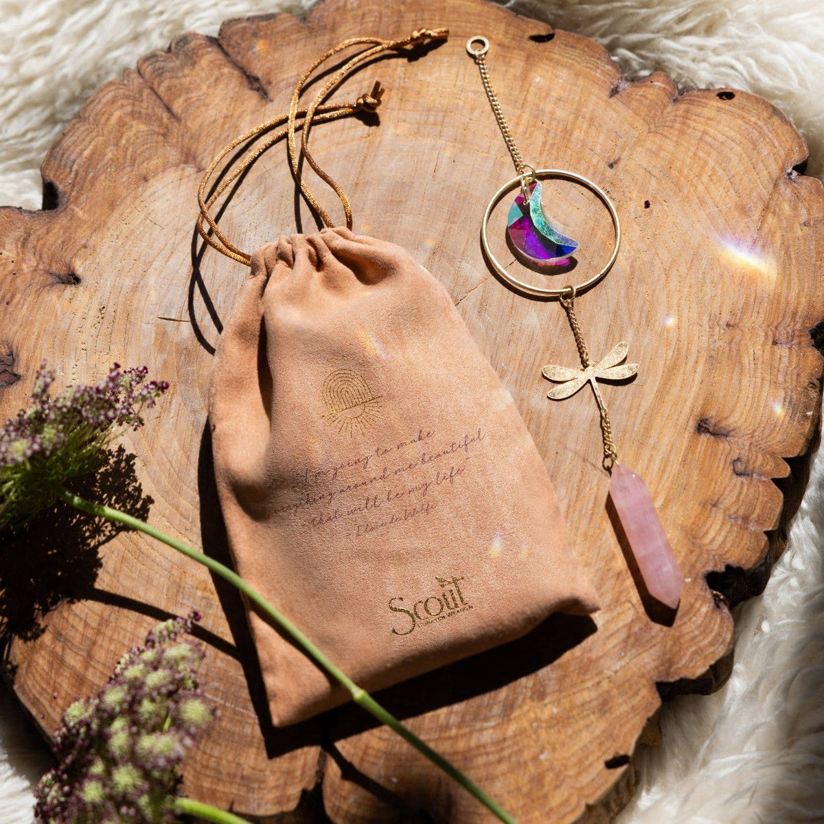 A tan drawstring bag and a gold bracelet featuring a dragonfly charm, pink crystal, and crescent moons rest on a wooden slab beside the Scout Curated Wears Stone Suncatcher - Dragonfly/Rose Quartz. Flowers and sunlight illuminate the enchanting scene.