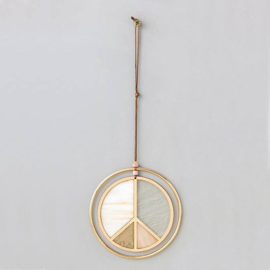 The Stained Glass Light Catcher - Peace Sign by Scout Curated Wears features a gold metal peace sign adorned with colored stone segments, suspended from a brown cord against a plain light gray wall—creating a striking effect reminiscent of stained glass art.