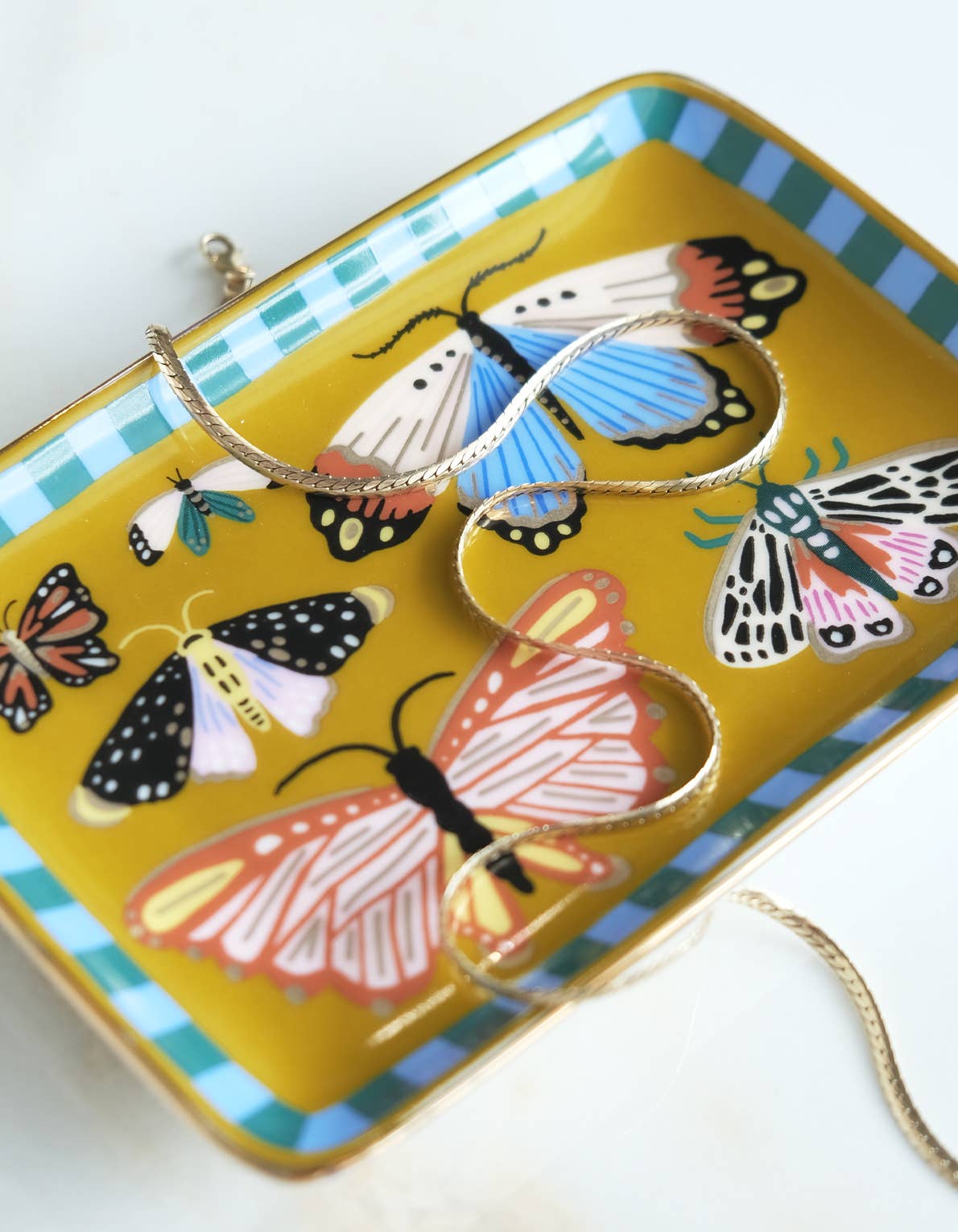 Idlewild Co. Porcelain Trinket Tray - Butterflies: a rectangular mustard-yellow ceramic dish with a blue and white striped rim, decorated with colorful illustrated butterflies and holding a thin gold chain necklace with gold detailing.