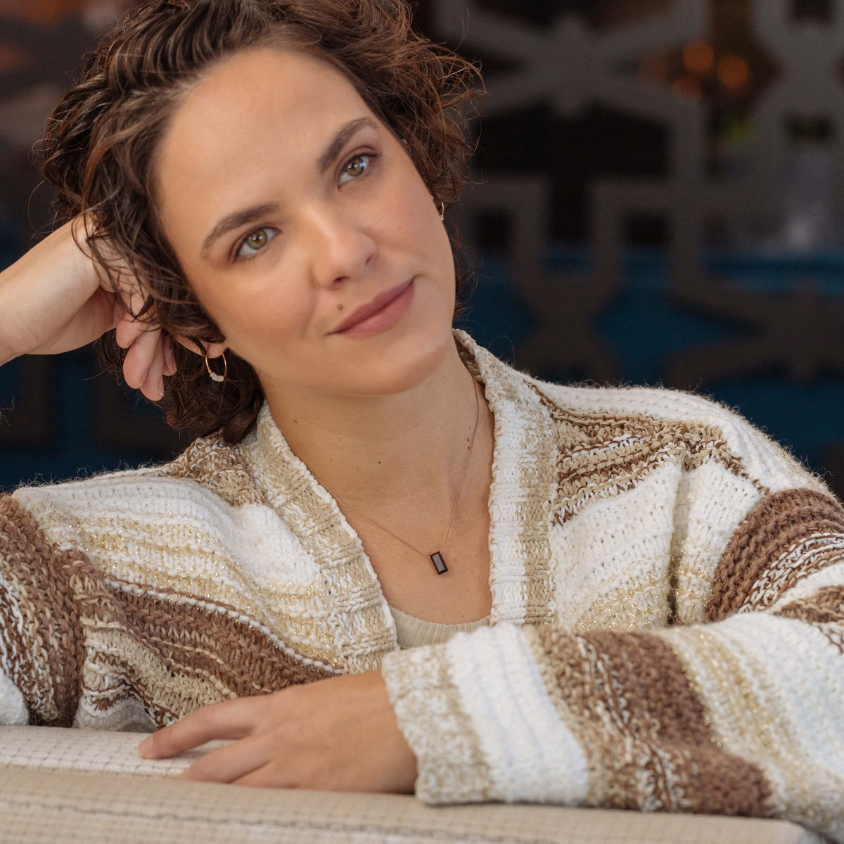 A person with short brown hair and hazel eyes leans on their hand, wearing a striped knit sweater and the SoulKu Smoky Quartz Refined Necklace for Release, looking thoughtful.
