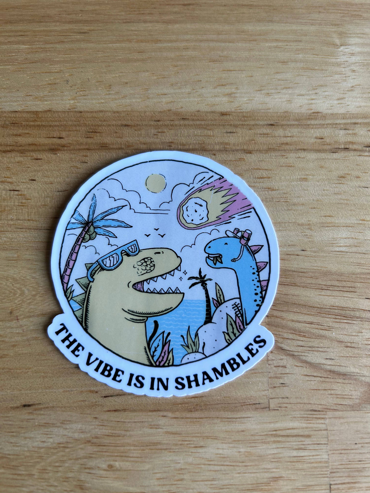 The Vibe is in Shambles Dinosaur Sticker by Lemonsgrace Designs features two cartoon dinos near water as a meteor falls—one sporting sunglasses. A fun way to show when the vibe is off!.