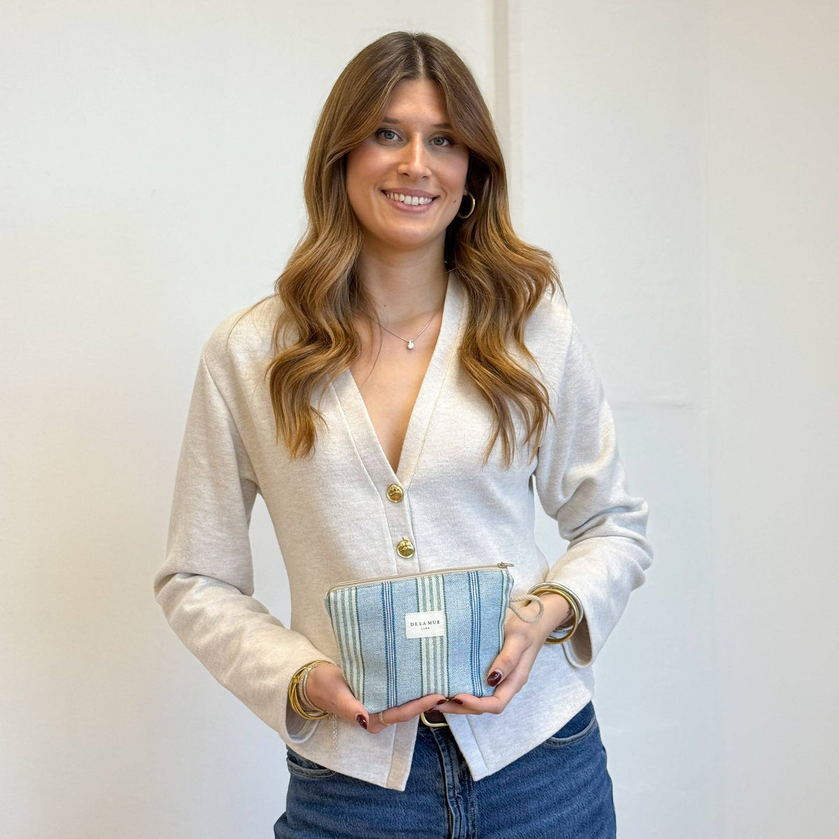 A woman with long brown hair, dressed in a light cardigan and jeans, smiles while holding the Striped Jacquard Small Makeup Bag – Blue & Green (Cata) by DE LA MUR LINE US Inc, featuring blue-striped jacquard fabric with a white label and made in Barcelona, against a plain white wall.