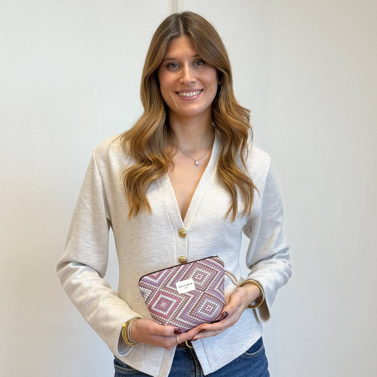A woman with long brown hair, dressed in a cream cardigan and jeans, stands smiling while holding the DE LA MUR LINE US Inc Geometric Jacquard Small Makeup Bag – Pink & Lilac (Nana) against a plain light background.
