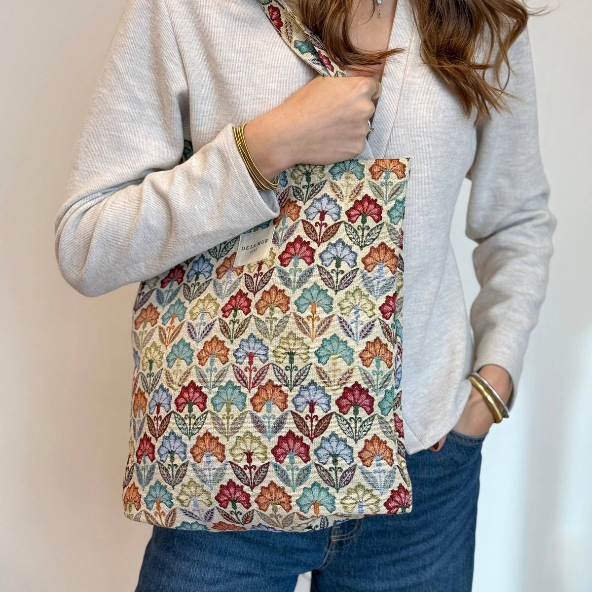 A person dressed in a light sweater and jeans carries the DE LA MUR LINE US Inc Floral Jacquard Medium Zippered Tote Bag – Multicolor (Flora), featuring a vibrant leaf-patterned design. Made in Barcelona, the tote hangs from their shoulder while they keep one hand in their pocket.