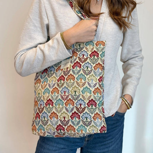 A person dressed in a light sweater and jeans carries the DE LA MUR LINE US Inc Floral Jacquard Medium Zippered Tote Bag – Multicolor (Flora), featuring a vibrant leaf-patterned design. Made in Barcelona, the tote hangs from their shoulder while they keep one hand in their pocket.