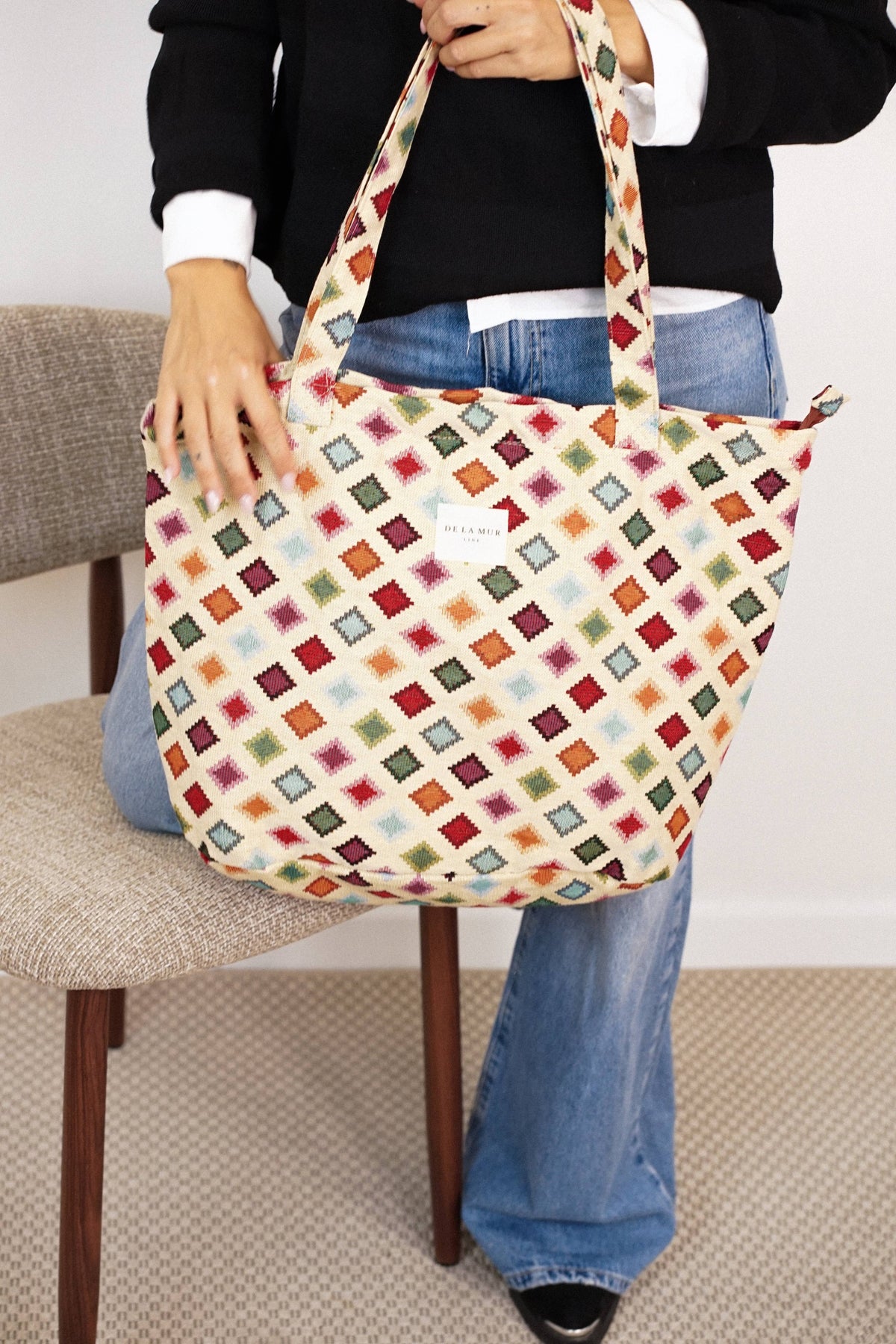 A person dressed in a black top and blue jeans sits on a chair, holding the Geometric Jacquard Large Zippered Tote Bag – Multicolor (Zac) by DE LA MUR LINE US Inc. The tote features a vibrant geometric jacquard pattern, two handles, and a small white label on the front.